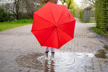 mischievous 5-year-old girl in rubber boots with red umbrella stands in rain puddle, capturing pure...