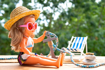Doll in bathing suit, summer straw hat, sunglasses, sea anchor, starfish on old wooden table,...