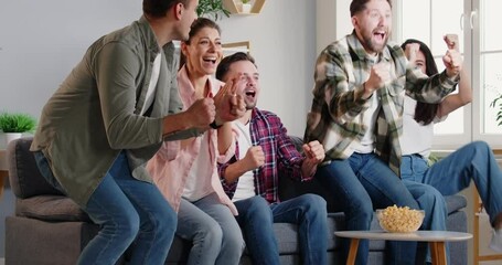Company of friends sitting on the sofa at home, watching a football or sport match on TV. They are eating popcorn, celebrating a goal, and supporting favorite soccer team together, enjoying the game.