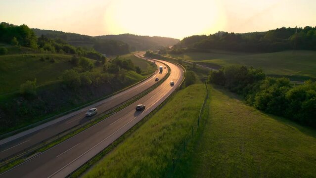 AERIAL, LENS FLARE: Winding highway glows in golden light of setting summer sun. Motorway with smooth flowing traffic winds past green fields and lush forests stretching across the hilly countryside.