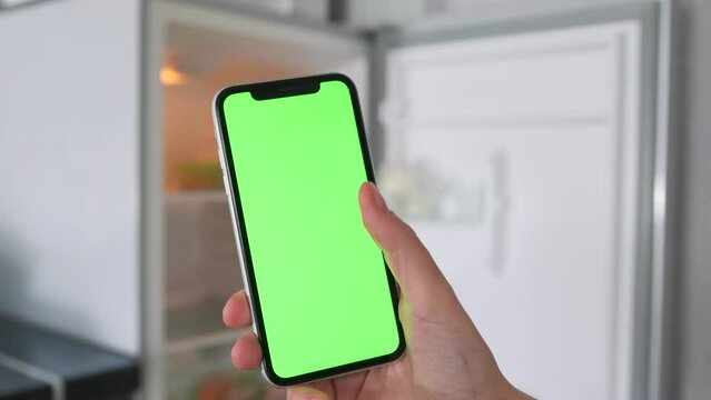 Woman's hand scrolls touch screen mobile phone with green chromakey against background of an open empty refrigerator, making purchases, looking at prices in app.