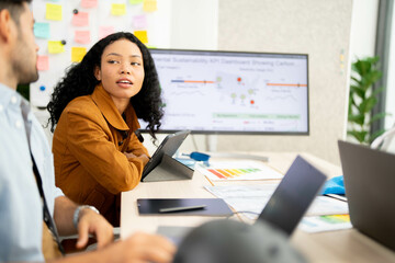Professional woman discusses data on a sustainability KPI dashboard during a collaborative office...