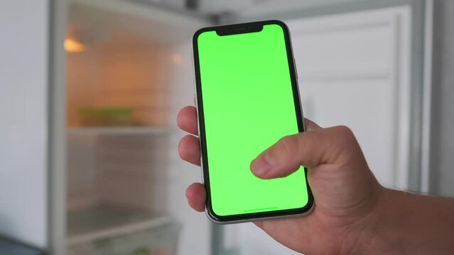 Close up hand holding smart phone using green screen, scrolling display, on empty shelves refrigerator blurred background.
