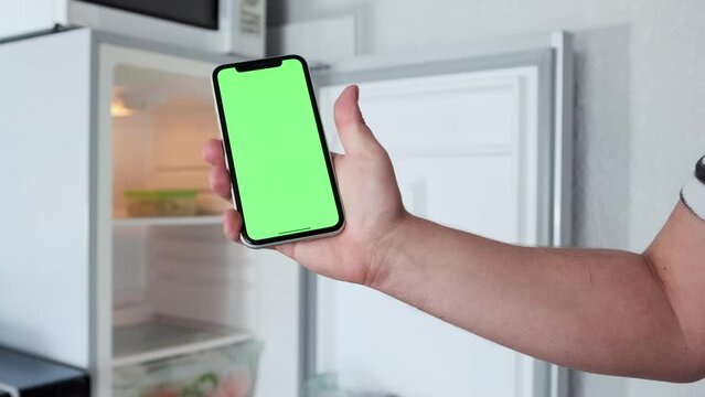 Mobile phone with green screen display in hand, by scrolling finger to touch screen, against the background of an open empty refrigerator, in kitchen.