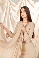 Young woman with long brunette hair dressed in a beige summer outfit poses confidently while holding a beige scarf.