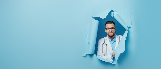 Content male doctor with glasses and a stethoscope breaks through a blue paper background, looking satisfied