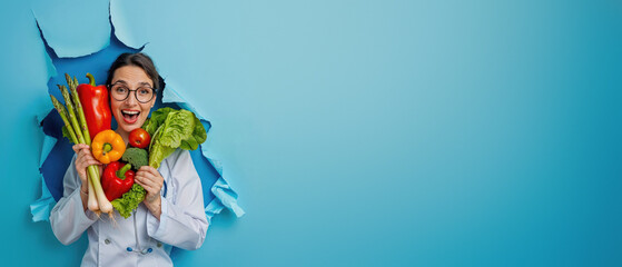 A smiling chef in uniform breaks through a blue background holding colorful fresh vegetables, portraying healthy eating