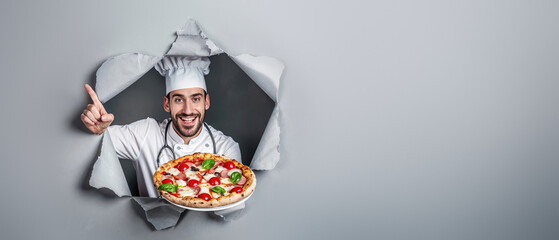 A chef points upwards and holds a pizza while peeking through a tear in grey paper