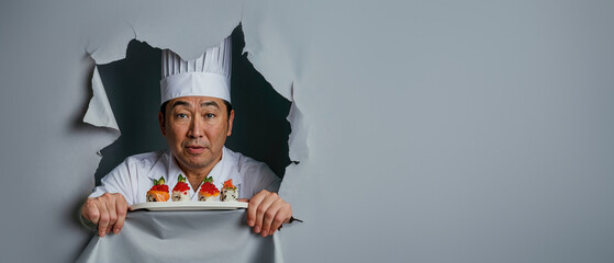 A professional Asian chef holds a sushi dish and peers through a large paper tear, showcasing his culinary art