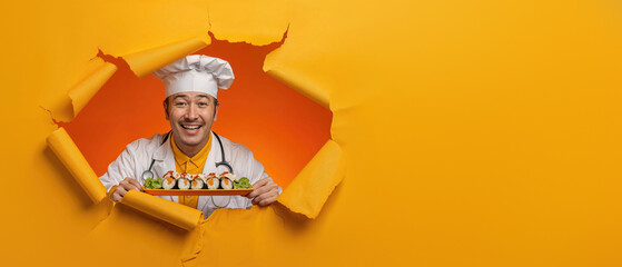 A joyful chef in white attire holds sushi while popping through a torn yellow paper background