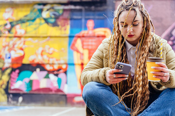 Thoughtful sad young hipster girl using smart phone against graffiti wall