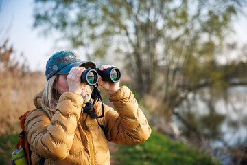 Woman tourist with binoculars looking for birds and animals at lake. Eco tourism. Joy of hiking in nature