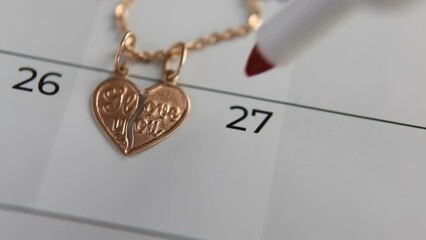 A gold pendant in the shape of a heart lies on the calendar date of the 27th. The memorable round wedding date is circled with a red marker. Two halves of a heart on a chain for lovers. Macro.