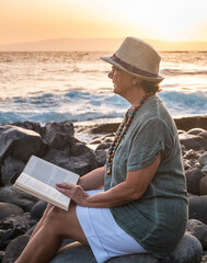 Side view of a senior woman alone sitting on the beach of pebbles with a book and looking the...