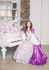 Beautiful woman in fantasy white and purple rococo style medieval dress sitting near piano with pink flowers
