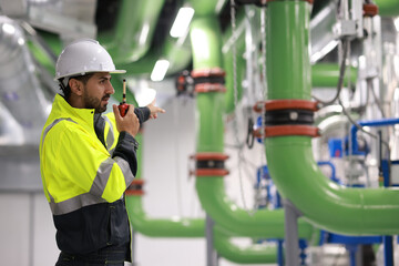 Mechanical engineer with green safety jacket holding laptop and walkie talkie working at site line of HVAC control room - obrazy, fototapety, plakaty