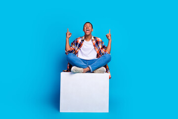 Full size photo of young man sit cube indicate fingers up empty space wear shirt isolated on blue...