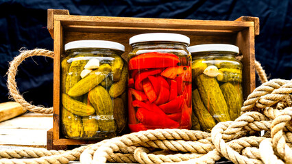 Glass jars with pickled red bell peppers and pickled cucumbers (pickles) isolated in wooden crate. Jars with variety of pickled vegetables. Preserved food concept in a rustic composition.
