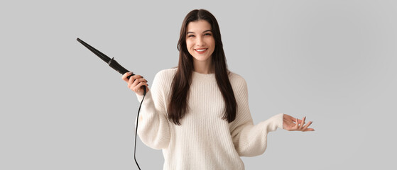 Beautiful young happy woman with curling iron on grey background