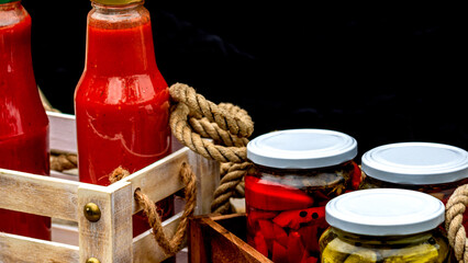 Wooden crate with glass jars with pickled red bell peppers and pickled cucumbers (pickles) isolated. Jars with variety of pickled vegetables. Preserved food concept in a rustic composition. - obrazy, fototapety, plakaty