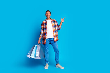Full size photo of nice young man hold bags point finger empty space wear shirt isolated on blue color background