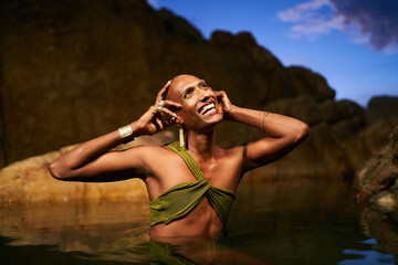Smiling gender fluid black person poses in natural still water pool. Queer ethnic fashion model in open dress, brass jewelry with gems standing gracefully in the middle of rocky lake at night. - Powered by Adobe