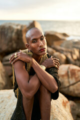 Non-binary ethnic fashion model in dress, brass jewelry sits on rocks by ocean. Trans sexual black...