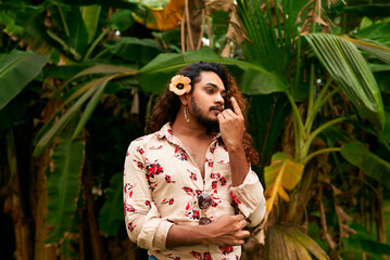 Sri Lankan man with flower in hair, stands in tropical garden, wearing floral shirt. Embodies gender fluidity, feminine expression, LGBT pride in natural setting. Poise reflects confidence, serenity.