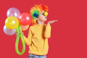 Funny little boy in clown costume with balloons blowing kiss on red background