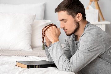 Young man with rosary beads and Bible praying in bedroom