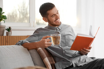 Young man with cup of tea reading book on sofa at home