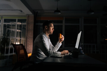 Side view of overworked businessman eating burger working on computer sitting on desk at office...