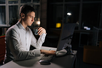 Side view of focused male reading on laptop working on deadline during late shift. Business, online and corporate worker drinking coffee and doing research on computer during overtime at work.