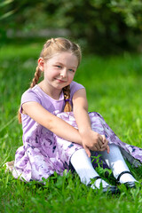 blondes glückliches Mädchen in einem lila Kleid sitz auf dem grünen Gras, schaut in die Kamera,...