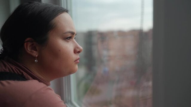 Distressed young adult female gazes from window, feeling isolated, pondering breakup. Solitude embraces her in urban setting, reflecting on lost love, battling silent depression.