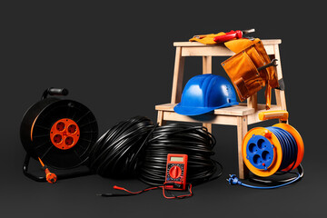 Stepladder with hardhat, rolled wires, extension cable reels and multimeter on black background