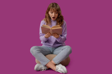 Surprised young woman reading book on purple background