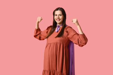 Beautiful young pregnant woman in superhero cape showing muscles on pink background