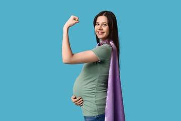 Beautiful young pregnant woman in superhero cape showing muscles on blue background