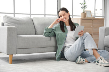 Pretty young woman with tablet computer sitting on floor near grey sofa in living room