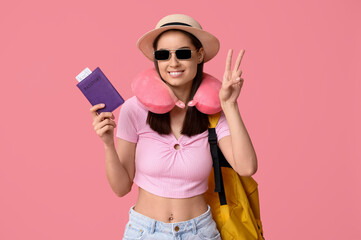 Young happy woman in travel pillow and sunglasses with passport and backpack showing peace gesture on pink background. Travel concept