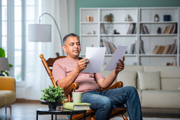 Indian mid age man with grey hair checking financial documents at home while sitting on rocking...