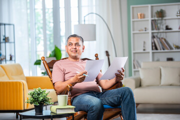 Indian mid age man with grey hair checking financial documents at home while sitting on rocking...