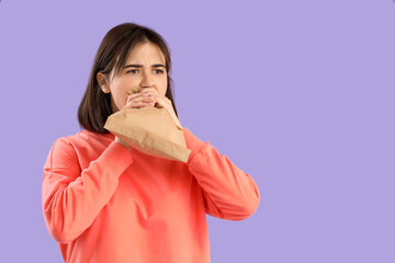 Beautiful young stressed woman with paper bag having panic attack on purple background. Stress Awareness Month - obrazy, fototapety, plakaty