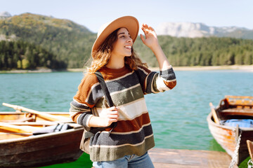 Traveling woman with a backpack and a hat enjoys the view of the mountain lake in sunny weather. The concept of travel, vacation. Active lifestyle.