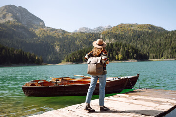 Traveling woman with a backpack and a hat enjoys the view of the mountain lake in sunny weather. The concept of travel, vacation. Active lifestyle.