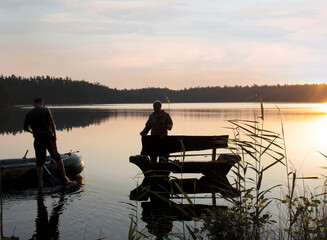 A man sits on a wooden bench and catches fish with a fishing rod. The second catches while standing. There is a beautiful forest lake around
