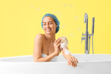 Young woman in shower cap with loofah taking bath on yellow background