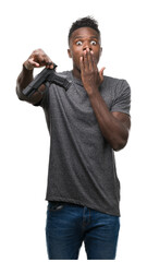 Young african american man holding a gun cover mouth with hand shocked with shame for mistake,...