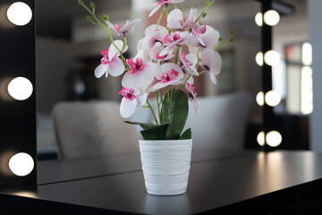 House furniture. Cozy interior. Delicate flower in white pot on dressing table with mirror and soft lighting